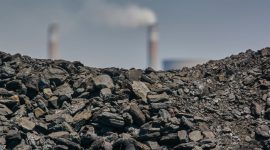 Tumpukan batu bara terlihat di depan cerobong asap sebuah fasilitas industri dengan latar belakang langit biru cerah. (Sumber: Bloomberg – Waldo Swiegers)