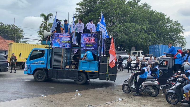 Massa buruh menggelar aksi unjuk rasa dengan mobil komando di ruas jalan Serang, membawa spanduk tuntutan penetapan UMK dan UMSK 2026, di bawah pengawalan aparat keamanan.