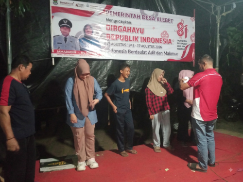 Kepala Desa Klebet Jamarudin mengenakan kemeja batik, tersenyum cerah saat menyerahkan piala besar dan piagam penghargaan kepada seorang wanita muda pemenang lomba karaoke di panggung panggung gembira perayaan HUT RI ke-81 di Kp. Santri Kepuh, Desa Klebet.